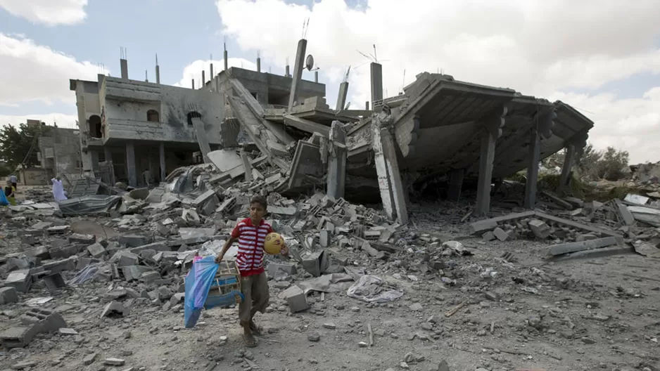 Un ni&ntilde;o palestino carga una pelota y una jaula de p&aacute;jaro mientras camina por los escombros de un edificio cerca al centro de refugiados de Rafah, al sur de Gaza. (Foto: AFP)